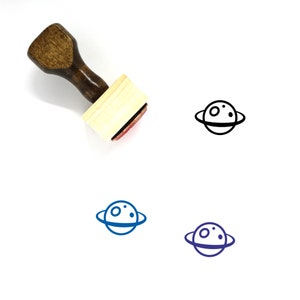 May include: A wooden rubber stamp with a black and white illustration of a planet with rings. The stamp is on a white background.