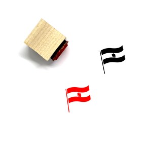 Puede incluir: Un sello de goma de madera con una almohadilla de tinta roja. El sello presenta una ilustraci&oacute;n en blanco y negro de una bandera con un s&iacute;mbolo. Tambi&eacute;n hay una bandera roja y blanca con un s&iacute;mbolo.