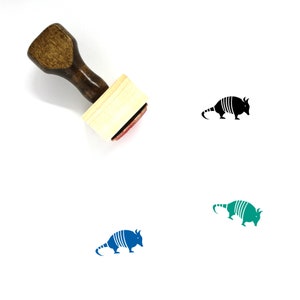 May include: A wooden rubber stamp with a handle and a rectangular base. The stamp features a black and white illustration of an armadillo. The stamp is shown on a white background.