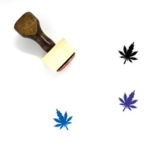 May include: A wooden rubber stamp with a handle and a rectangular base. The stamp features a black and white marijuana leaf design. There are two other marijuana leaf designs in the image, one in blue and one in dark blue.