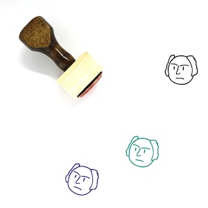 May include: A wooden rubber stamp with a brown handle and a rectangular wooden block. The stamp features a black line drawing of a person's face with a serious expression.