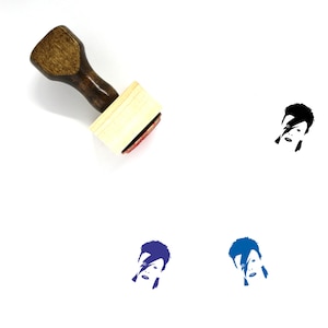 May include: A wooden rubber stamp with a black and white image of David Bowie's face. The stamp is on a white background. There are three other images of David Bowie's face in different colors below the stamp.