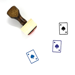 Puede incluir: Un sello de goma de madera con un mango marrón y una base rectangular de madera. El sello presenta una ilustración en blanco y negro de una carta de juego con el as de picas.