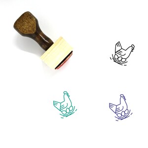 Puede incluir: Un sello de goma de madera con un mango marrón y una base rectangular. El sello presenta un dibujo lineal de una gallina sentada en un nido de huevos.