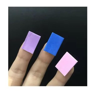 Puede incluir: Tres piezas rectangulares de arte de u&ntilde;as en lavanda, azul y rosa se muestran en los dedos sobre un fondo negro. Las piezas de arte de u&ntilde;as tienen forma cuadrada y parecen estar hechas de un material s&oacute;lido.