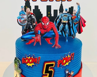 May include: A blue and red superhero-themed birthday cake. The cake features a blue base with superhero figures, including Spider-Man, Captain America, Thor, Batman, and Superman. The cake is decorated with the name "Kylian Kai" and the number "5".