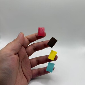 Puede incluir: Una mano sosteniendo cinco peque&ntilde;os bloques rectangulares en rojo, marr&oacute;n, amarillo y azul. Los bloques parecen ser de madera o un material similar, y est&aacute;n espaciados uniformemente en los dedos.