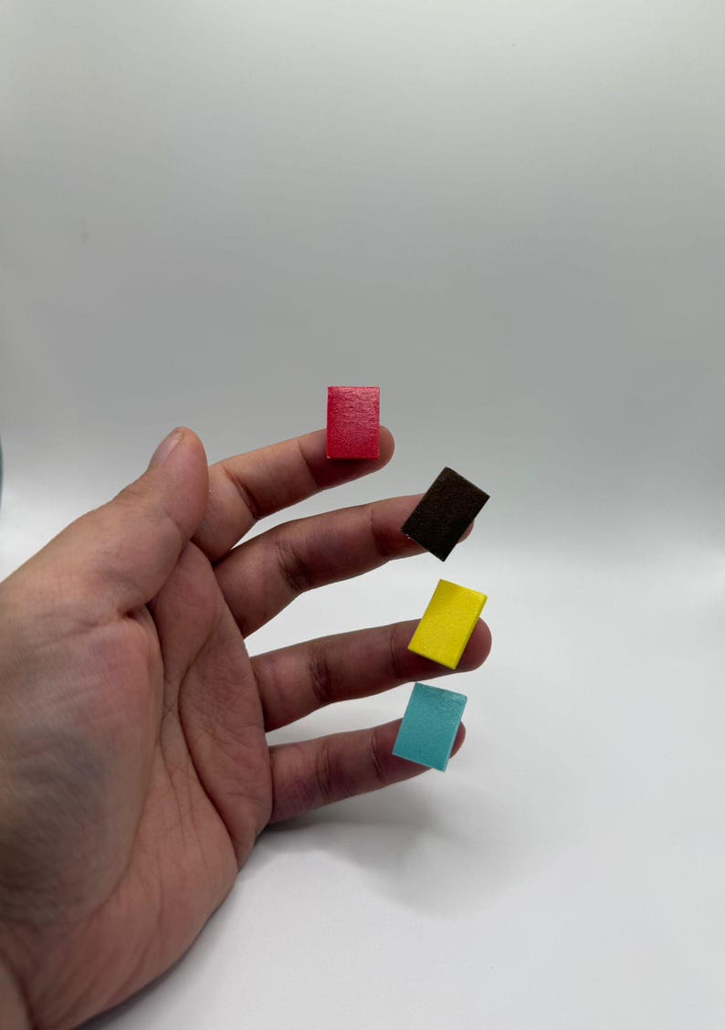 Puede incluir: Una mano sosteniendo cinco peque&ntilde;os bloques rectangulares en rojo, marr&oacute;n, amarillo y azul. Los bloques parecen ser de madera o un material similar, y est&aacute;n espaciados uniformemente en los dedos.