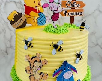 May include: A yellow Winnie the Pooh themed cake decorated with Winnie the Pooh, Piglet, Tigger, and Eeyore characters. The cake is decorated with a honey pot, bees, and a sign that says "Chico 4 meses".