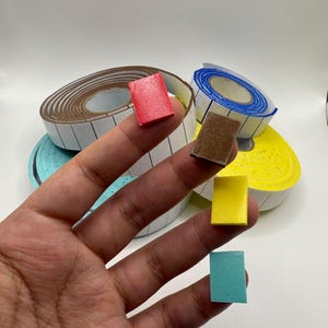 Puede incluir: Una mano sosteniendo peque&ntilde;os bloques rectangulares de colores: rojo, marr&oacute;n, amarillo y turquesa. En el fondo, rollos de cinta de etiquetas blancas con bordes marrones y azules. Los bloques son probablemente para manualidades u organizaci&oacute;n.