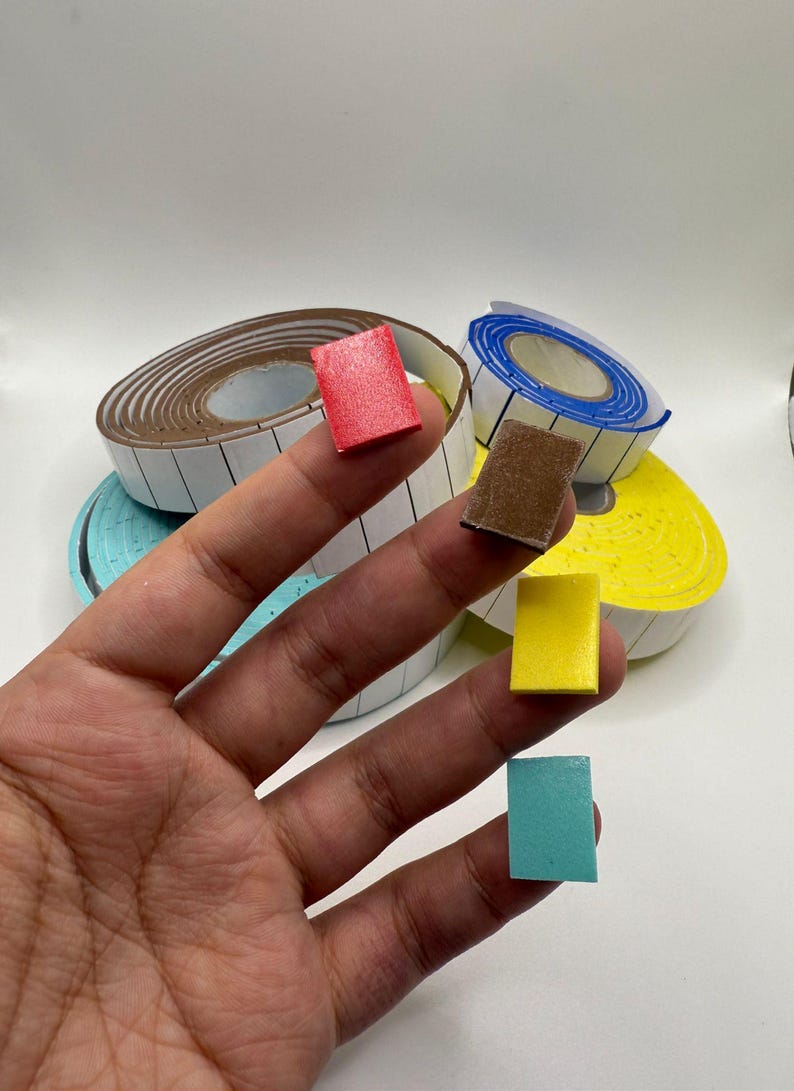 Puede incluir: Una mano sosteniendo peque&ntilde;os bloques rectangulares de colores: rojo, marr&oacute;n, amarillo y turquesa. En el fondo, rollos de cinta de etiquetas blancas con bordes marrones y azules. Los bloques son probablemente para manualidades u organizaci&oacute;n.