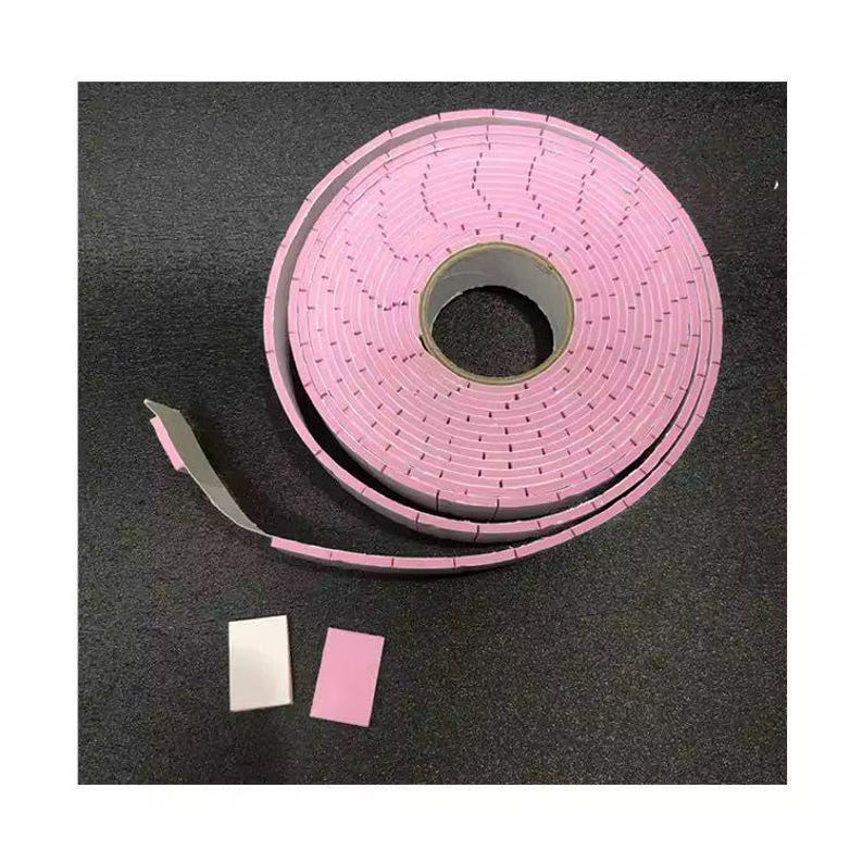 Puede incluir: Un rollo de cinta de espuma rosa con respaldo de papel, desenrollado sobre una superficie oscura. Dos peque&ntilde;os trozos rectangulares de la cinta est&aacute;n separados del rollo. La cinta est&aacute; dise&ntilde;ada con perforaciones espaciadas.