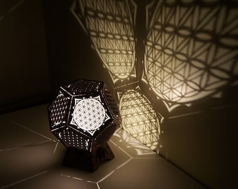 May include: A wooden lamp with a geometric design casts intricate shadows on a white surface. The lamp is shaped like a dodecahedron and features a flower of life pattern.