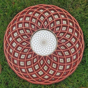 Op de afbeelding: Een ronde houten wanddecoratie met een rood en zwart geometrisch patroon. Het midden van het ontwerp heeft een wit en gouden mandala.