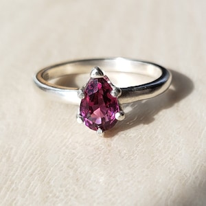 May include: A silver ring with a pear-shaped, deep pink gemstone set in a prong setting.