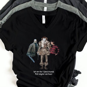 以下が含まれることがあります： 黒、チャコール、グレーのVネックTシャツのスタック。前面の黒いシャツには、3人のホラー映画のキャラクターのグラフィックと「We are the 3 best friends that anyone can have」というテキストが表示されています。