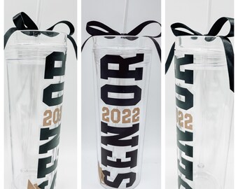 May include: Three clear plastic tumblers with black lids and black straws. Each tumbler has a black vinyl decal that says "SENIORS 2022" in black with a gold outline. Each tumbler has a black ribbon tied around the lid in a bow.