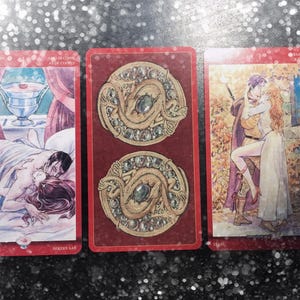 May include: Three rectangular tarot cards with red borders. The left card shows a couple embracing in bed with a silver chalice above. The center card features two circular gold medallions with dragons and green gems. The right card depicts a couple kissing.