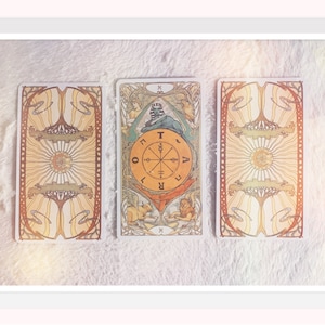Puede incluir: Tres cartas de tarot colocadas sobre una superficie blanca y peluda. La carta del medio es la carta de la Rueda de la Fortuna, que muestra una rueda con las letras 'T', 'O', 'R', 'A', 'V', 'E', 'L' e 'I' alrededor de la rueda. La carta est&aacute; rodeada de un dise&ntilde;o colorido con una esfinge y un le&oacute;n.