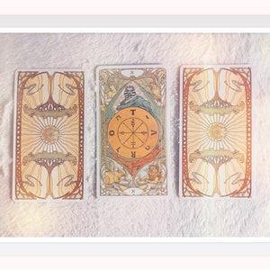 Op de afbeelding: Drie tarotkaarten die op een wit, pluche oppervlak liggen. De middelste kaart is de kaart van het Rad van Fortuin, die een wiel met de letters 'T', 'O', 'R', 'A', 'V', 'E', 'L' en 'I' rond het wiel toont. De kaart is omgeven door een kleurrijk ontwerp met een sfinx en twee figuren.