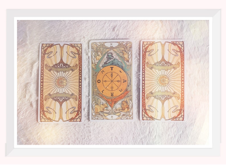 Op de afbeelding: Drie tarotkaarten die op een wit, pluche oppervlak liggen. De middelste kaart is de kaart van het Rad van Fortuin, die een wiel met de letters 'T', 'O', 'R', 'A', 'V', 'E', 'L' en 'I' rond het wiel toont. De kaart is omgeven door een kleurrijk ontwerp met een sfinx en twee figuren.