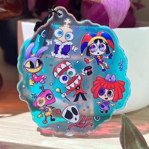 Puede incluir: Un dije circular de acrílico iridiscente que presenta una colección de coloridos personajes de dibujos animados. El diseño incluye un conejo morado, un bufón, una figura con sombrero rojo y bastón, una muñeca de pelo rojo y una máscara blanca sobre un fondo verde azulado.