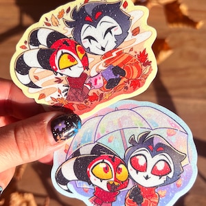 May include: Two cartoon stickers featuring characters with red, black, and white designs. One sticker shows the characters with autumn leaves, and the other shows them under an umbrella. The stickers have a glittery finish.