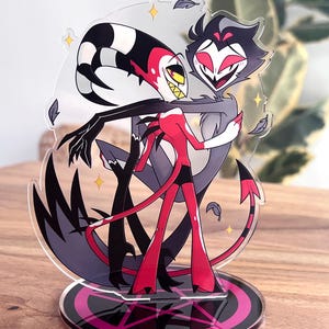 May include: A clear acrylic stand featuring two cartoon characters embracing. One character is red and black with a tail, the other is gray. The stand has a black and pink base with a star design.