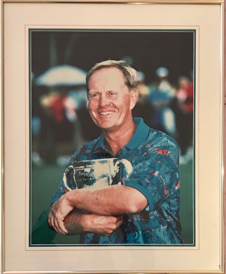 May include: A man in a blue and red patterned shirt smiles while holding a silver trophy. He is standing in front of a blurred background of people.