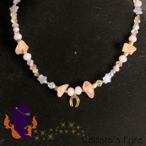 Peut inclure: Un collier de perles avec un pendentif en fer à cheval doré, des perles roses et blanches, et des étoiles. Le collier est présenté sur un fond sombre. Le texte "Callisto's Fyre" est visible.