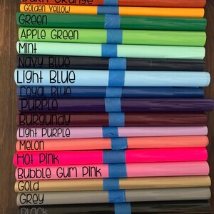 以下が含まれることがあります： 赤、オレンジ、黄色、緑、青、紫、ピンク、金、グレー、黒、白など、さまざまな色のロール状ビニールシートのコレクション。各ロールには、黒いテキストで色名が記載されており、青いテープで固定されています。