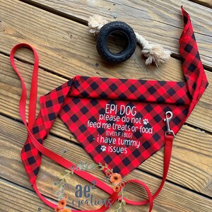 以下が含まれることがあります： 「EPI DOG please do not feed me treats or food (EVEN IF I BEG) - I have tummy issues」のテキストが入った赤と黒のバッファローチェックの犬用バンダナ。赤いリードと黒いタイヤの犬用おもちゃも画像にあります。
