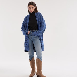 May include: A long-sleeved blue cardigan featuring a black geometric pattern, worn over a black turtleneck shirt, light blue denim jeans, and tan suede knee-high boots. The cardigan has an open front design with a black trim along the edges.
