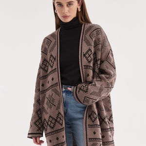 May include: A long-sleeved, open-front cardigan in a muted mauve color featuring a repeating black geometric pattern. The knit garment has a straight hemline and wide sleeves, worn over a black mock-neck top and light blue denim jeans.