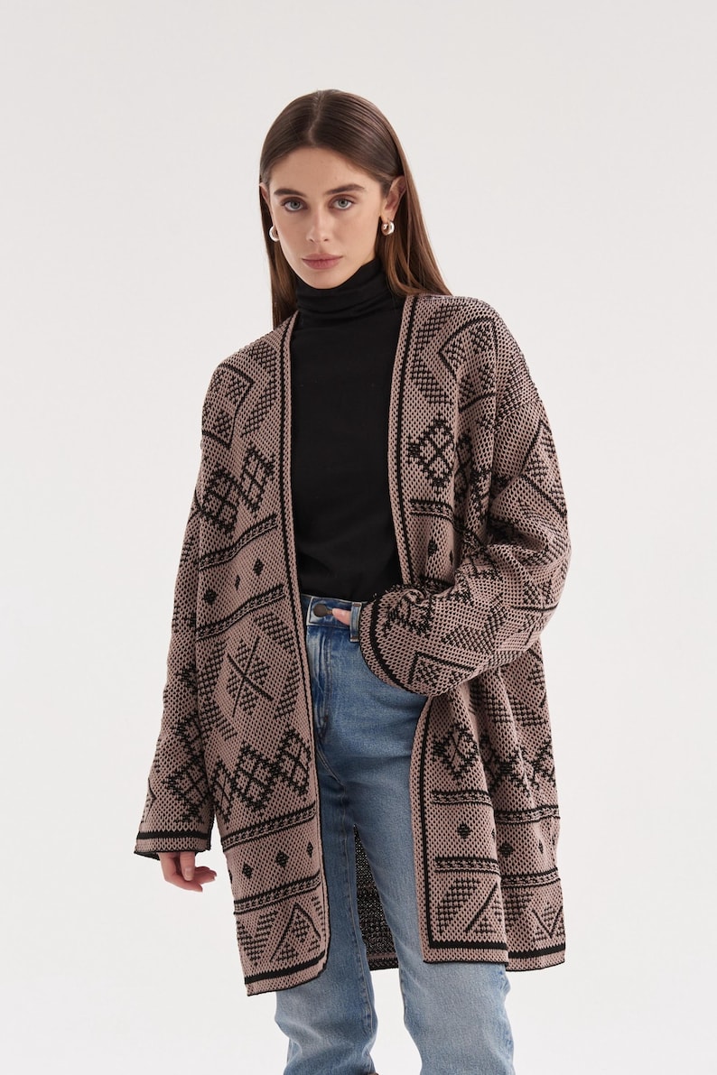 May include: A long-sleeved, open-front cardigan in a muted mauve color featuring a repeating black geometric pattern. The knit garment has a straight hemline and wide sleeves, worn over a black mock-neck top and light blue denim jeans.
