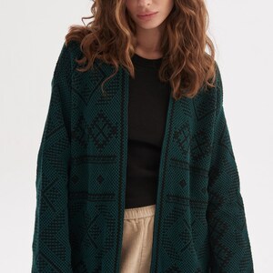 May include: A long-sleeved, open-front cardigan in a deep forest green hue, featuring a textured knit pattern with contrasting black geometric diamond and line motifs throughout the fabric, including a solid black trim along the front opening edges.