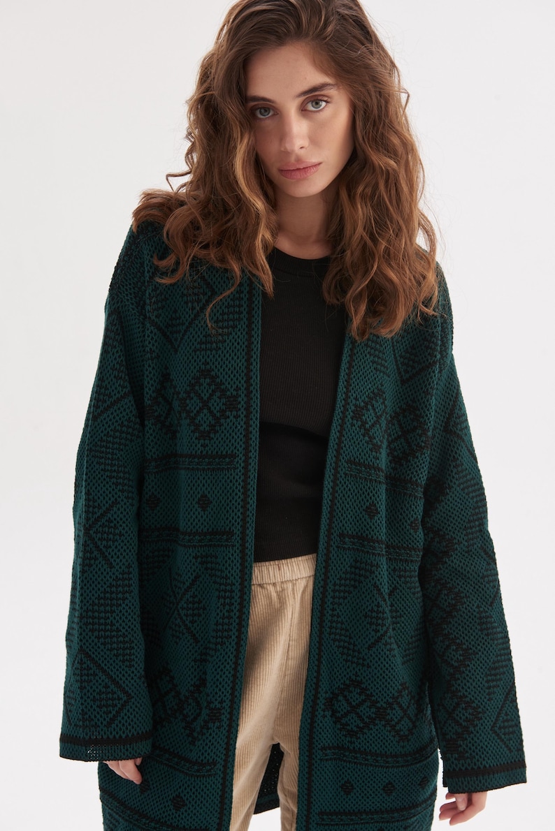 May include: A long-sleeved, open-front cardigan in a deep forest green hue, featuring a textured knit pattern with contrasting black geometric diamond and line motifs throughout the fabric, including a solid black trim along the front opening edges.