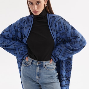 May include: An open-front knit cardigan featuring a vibrant blue and black geometric pattern, worn over a black turtleneck shirt and light blue denim jeans. The long-sleeved cardigan has a relaxed fit and a textured knit construction throughout.