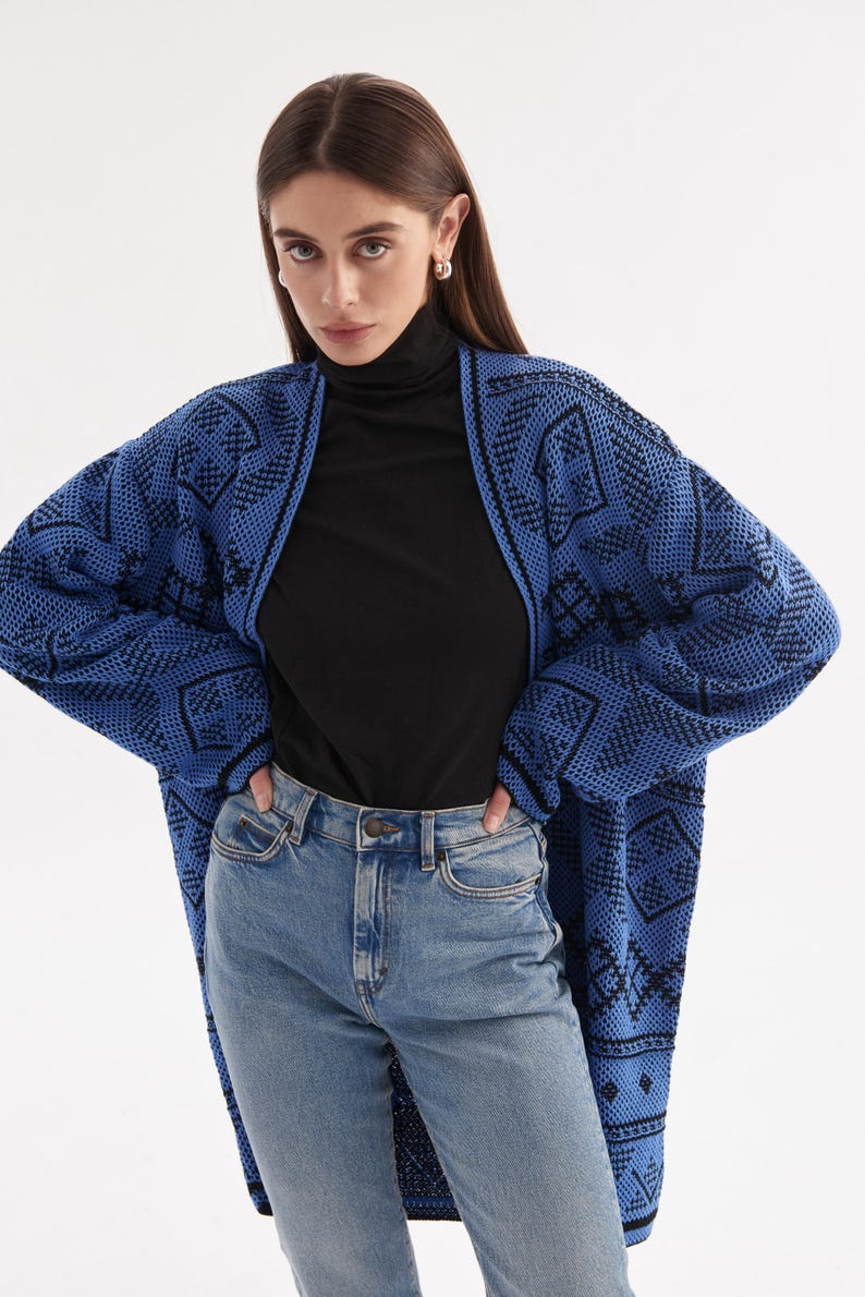 May include: An open-front knit cardigan featuring a vibrant blue and black geometric pattern, worn over a black turtleneck shirt and light blue denim jeans. The long-sleeved cardigan has a relaxed fit and a textured knit construction throughout.