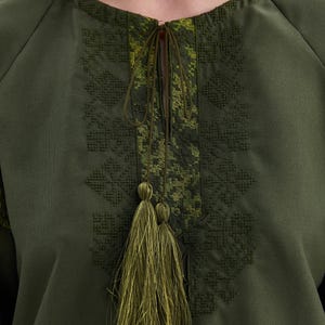 May include: Olive green tunic top with intricate embroidered detailing around the neckline and sleeves. The top features a drawstring closure and a decorative tassel. The embroidery is in shades of green, creating a textured and visually appealing design.