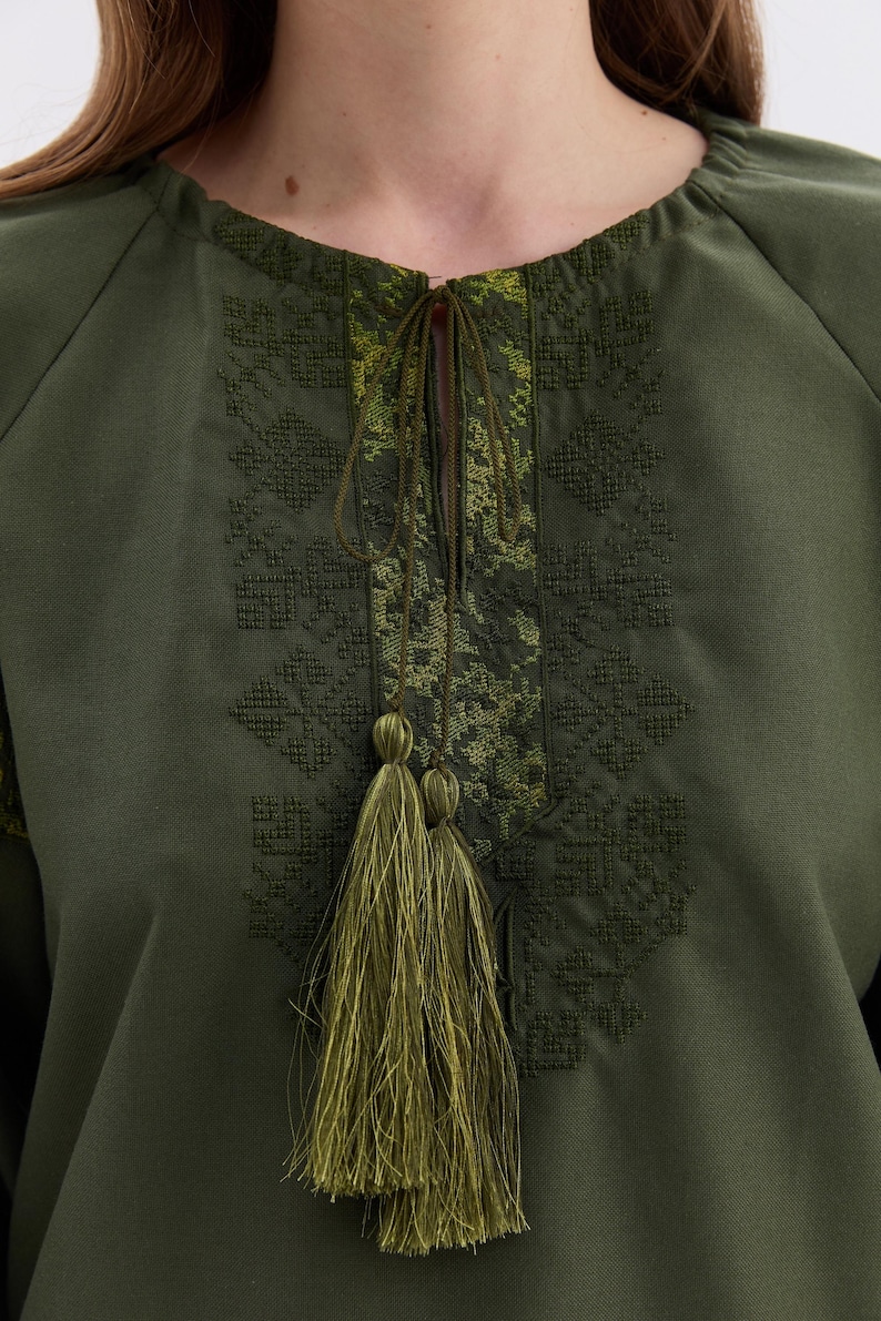 May include: Olive green tunic top with intricate embroidered detailing around the neckline and sleeves. The top features a drawstring closure and a decorative tassel. The embroidery is in shades of green, creating a textured and visually appealing design.