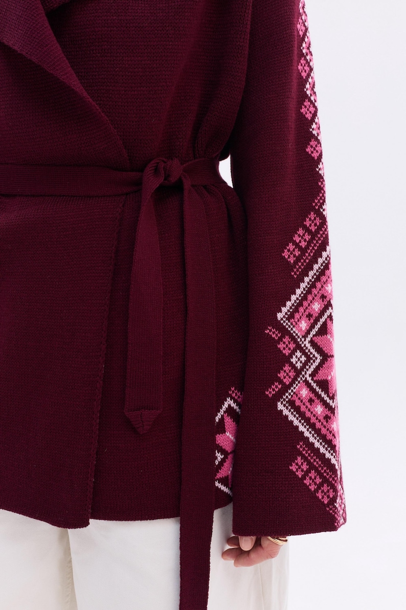 May include: A burgundy wrap cardigan with a matching tie belt. The sleeve features a geometric pattern in shades of pink and white. The cardigan is made of a soft knit material.