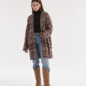 May include: A long-sleeved, open-front cardigan in a muted taupe color featuring a repeating black geometric pattern. The garment is worn over a black turtleneck sweater, light blue denim jeans, and tan suede knee-high western-style boots.