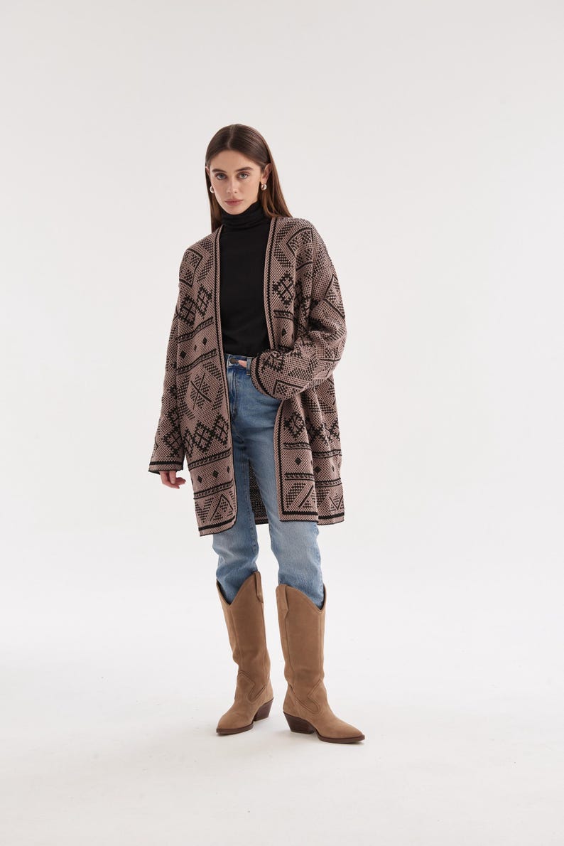 May include: A long-sleeved, open-front cardigan in a muted taupe color featuring a repeating black geometric pattern. The garment is worn over a black turtleneck sweater, light blue denim jeans, and tan suede knee-high western-style boots.