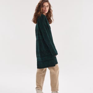 May include: A long-sleeved dark green knit sweater featuring a black geometric diamond pattern, paired with beige trousers and light-colored ankle boots. The sweater has a crew neckline and a straight hemline that reaches the mid-thigh area.