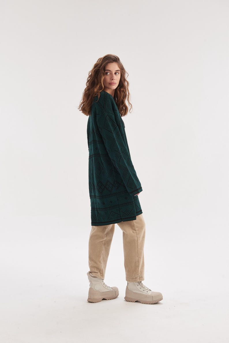 May include: A long-sleeved dark green knit sweater featuring a black geometric diamond pattern, paired with beige trousers and light-colored ankle boots. The sweater has a crew neckline and a straight hemline that reaches the mid-thigh area.
