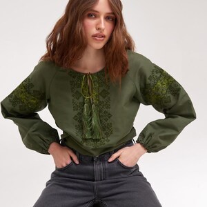 May include: Olive green long-sleeve blouse with intricate embroidered details on the sleeves and bodice. The blouse features a round neckline with a drawstring and tassel accents. The sleeves are gathered at the wrists. The model is wearing dark gray jeans.