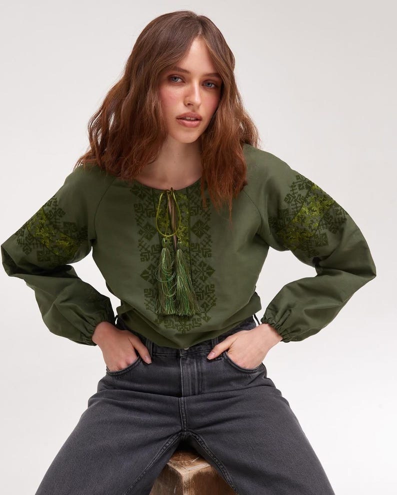 May include: Olive green long-sleeve blouse with intricate embroidered details on the sleeves and bodice. The blouse features a round neckline with a drawstring and tassel accents. The sleeves are gathered at the wrists. The model is wearing dark gray jeans.