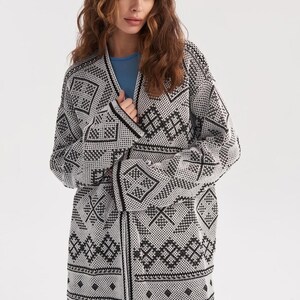May include: An open-front cardigan featuring a black and white geometric pattern throughout. The knit garment has long sleeves, a straight hemline, and a V-neckline, worn over a solid light blue shirt. The design consists of repeating diamond and linear motifs.