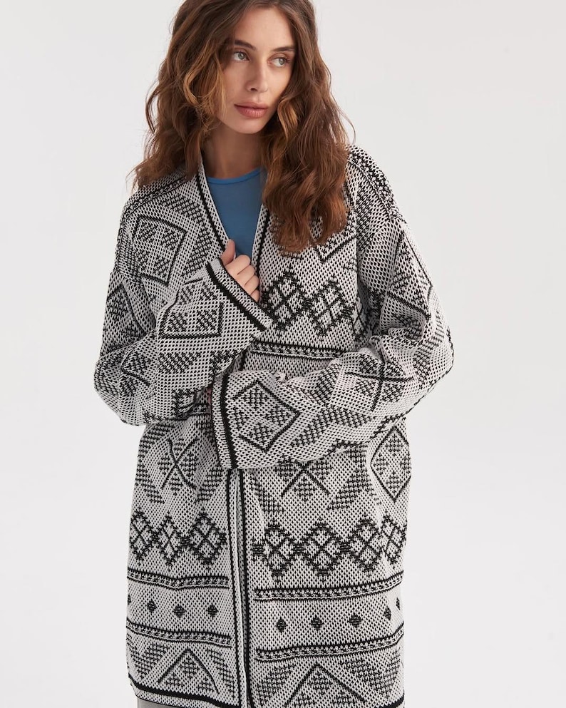 May include: An open-front cardigan featuring a black and white geometric pattern throughout. The knit garment has long sleeves, a straight hemline, and a V-neckline, worn over a solid light blue shirt. The design consists of repeating diamond and linear motifs.